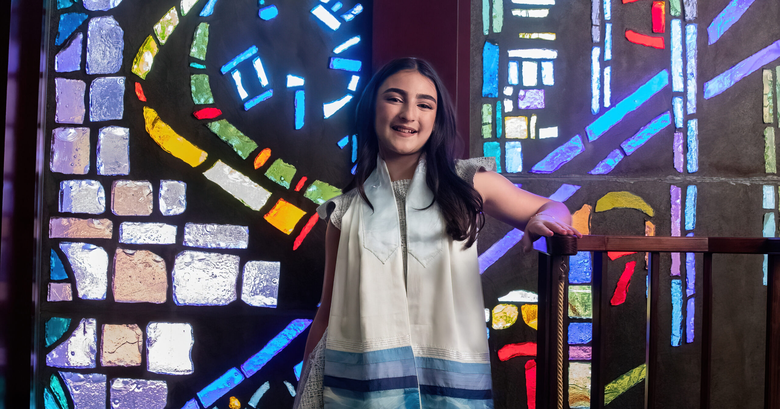 A Jewish teenage girl with brown hair poses in front of a stain glass window in a synagogue.