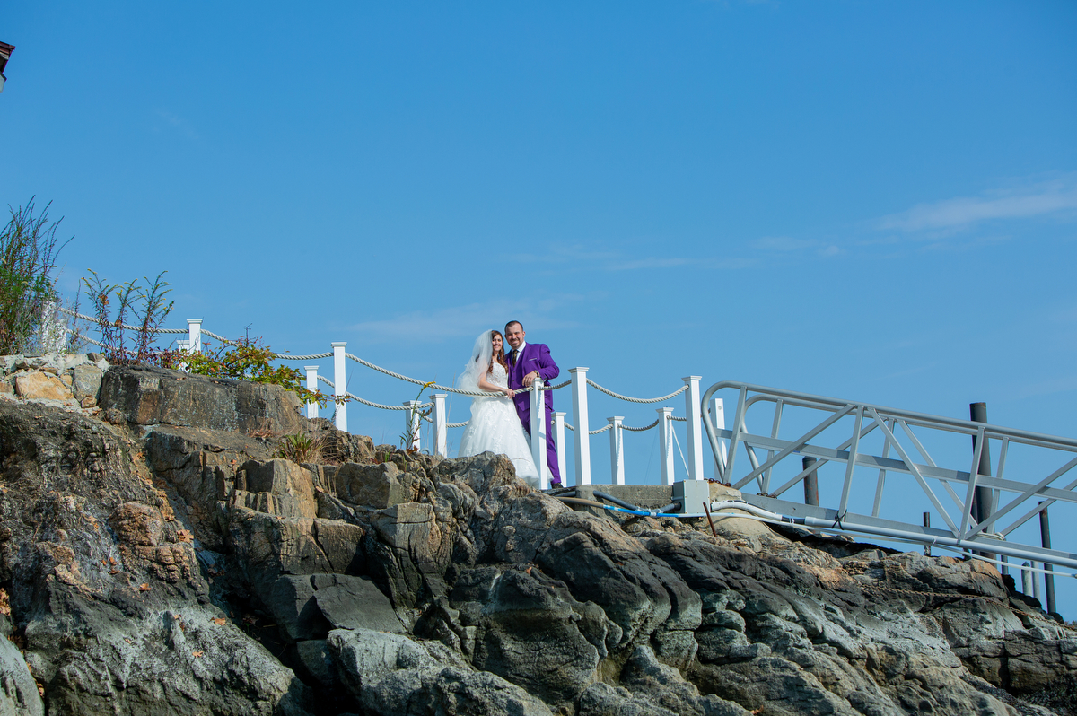 A groom in a purple suit kisses a bride in a white dress against a deep blue sky on the rocky shore of Long Island Sound Westcheter Wedding Photographer