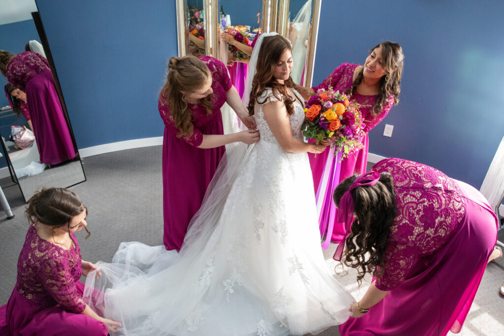 Bridesmaids in fuschia dresses help a bride in a white dress with finishing touches as she gets ready at the Mamaroneck Beach Club Westchester Wedding Photographer