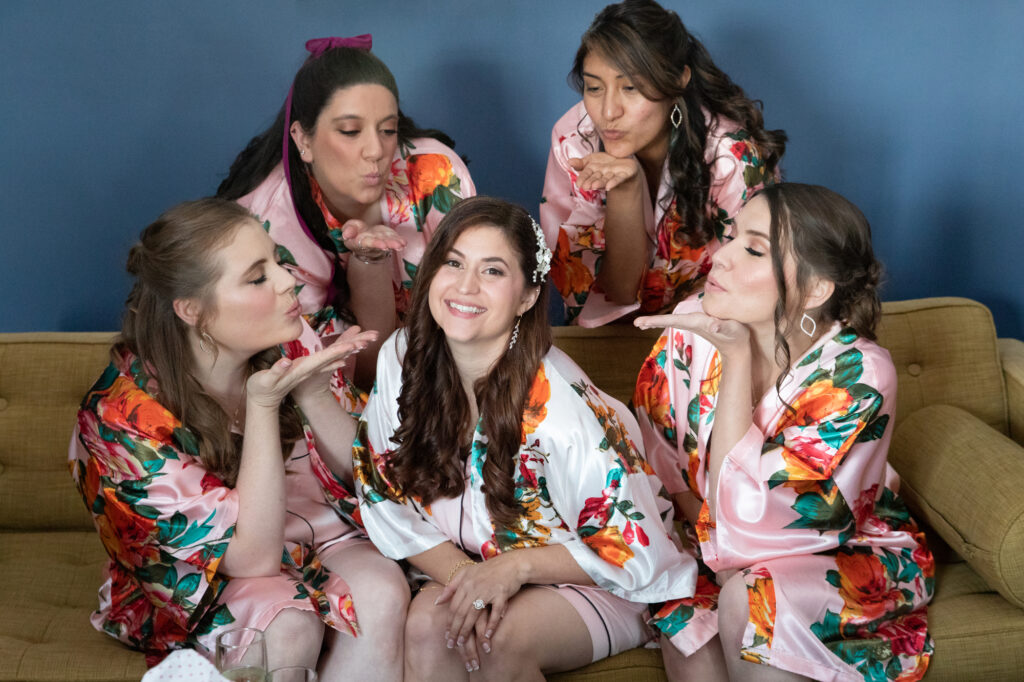 Bridesmaids in flowered robes blow a kiss to a bride in a white robe during getting ready photos at the Mamaroneck Beach Club Westchester Wedding Photographer