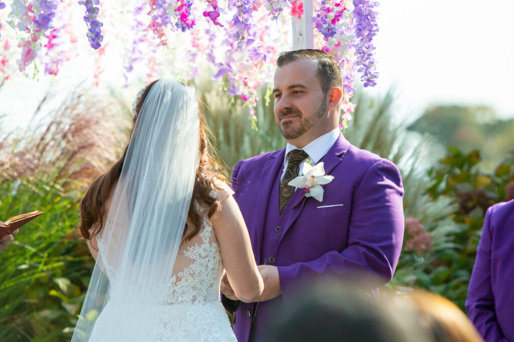 A bride with brown hair wearing a white veil faces a groom in a purple suit during vows at the Mamaroneck Beach Club Westchester Wedding Photographer