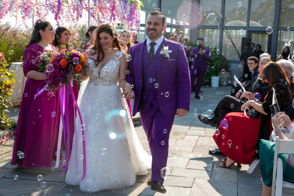A bride in a white dress and a groom i a pruple suit recess from their cereony in a cloud of bubbles provided by Magicl Memories entertainment Westchester Wedding Photographer