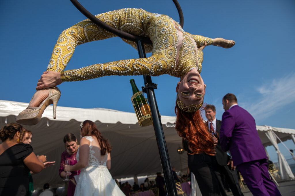 An acrobat with red hair in a gold leotard hired by Magical Memories Entertainment bends through a hoop during cocktail hour Westchester wedding Photographer Milestone Images