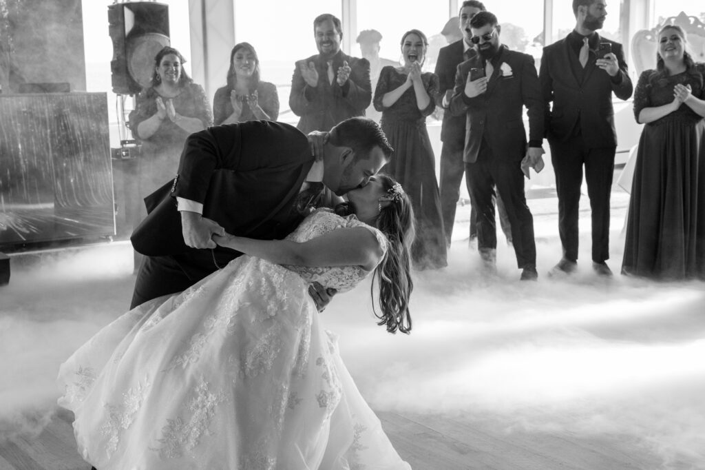A groom dips his bride during their first dance surrounded by fog from a fog machine and their cheering wedding party in a black and white photo at the Mamaroneck Beach Club Westchester Wedding Photographer