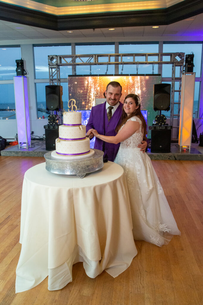 A groom in a purple suit and a bride in a white dress cut a three-tier wedding cake with white frosting and purple ribbon Westchester wedding Photographer Milestone Images