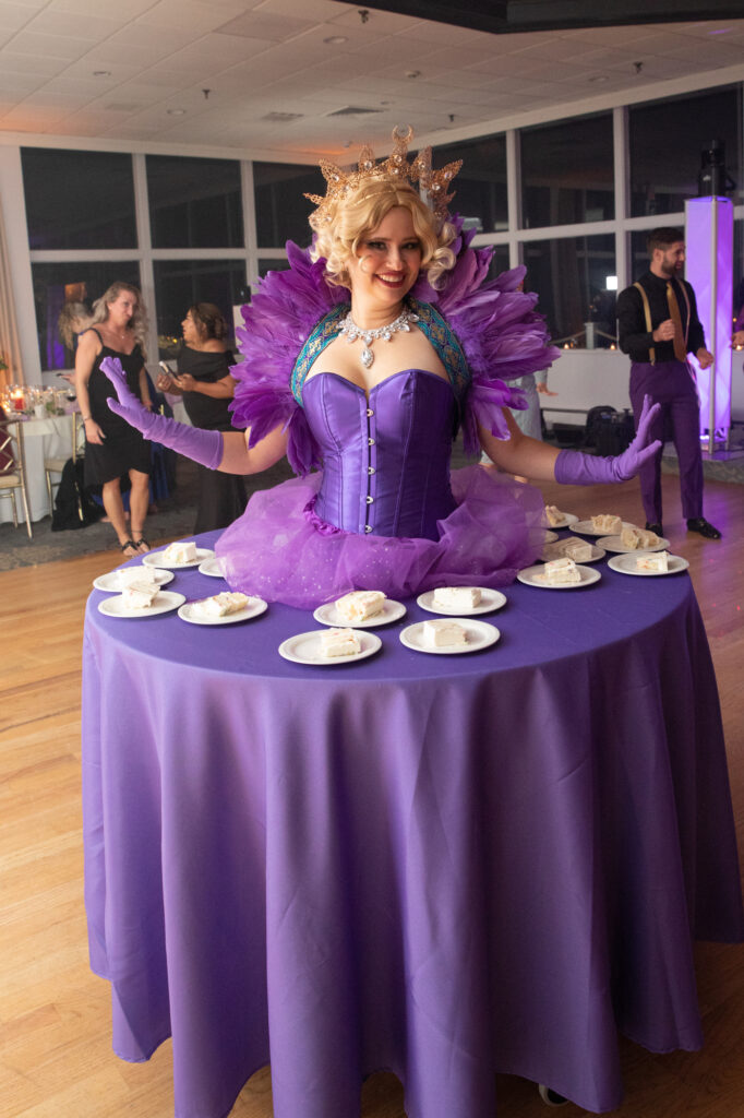 A blond female performer wears purple feathers and gloves as her purple tutu becomes a rolling table with slices of cake on it Westchester wedding Photographer Milestone Images