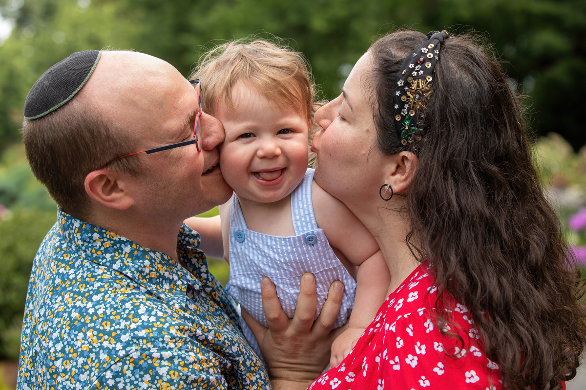 A father in a blue shirt and a mother in a red dress kiss their toddler on the cheeks during family photos in Fort Tryon Park New York Family Photographer Milestone Images