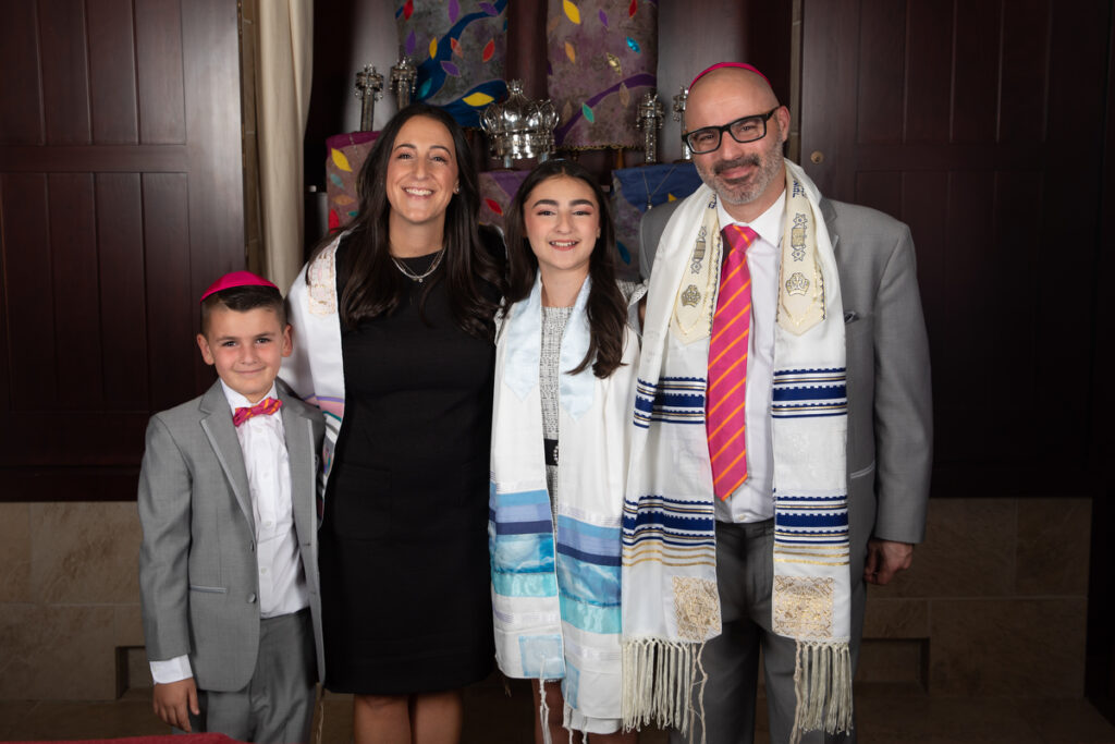 A family of four with a 7-year-old, mother, father and bat mitzvah girl wear coordinating outfits with pink and orange details in front of an open ark at Orangetown Jewish Center Rockland County Bat Mitzvah Photographer